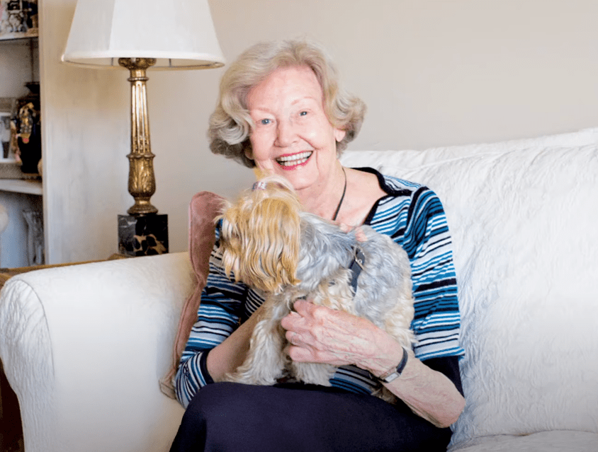 A smiling senior woman sits on a couch while holding a small dog