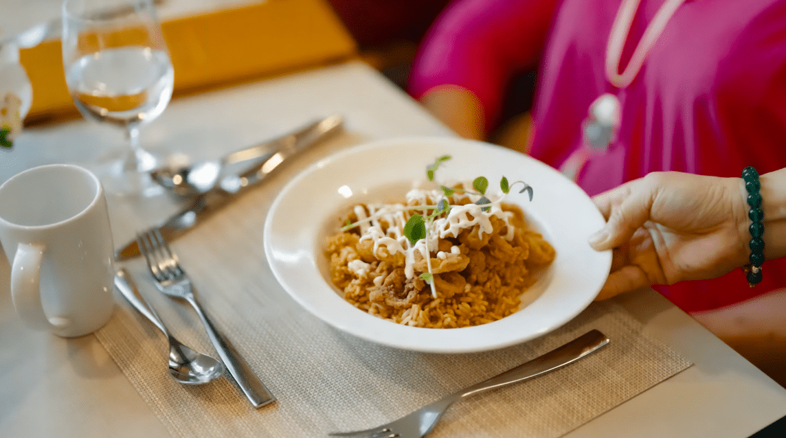 A plate of fancy pasta topped with white sauce and garnishes is placed on a set dining table, in front of a woman in a pink shirt.