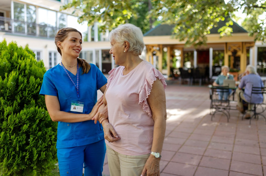 A staff member in a blue uniform smiles warmly while walking arm-in-arm with a senior resident outdoors at a retirement community.