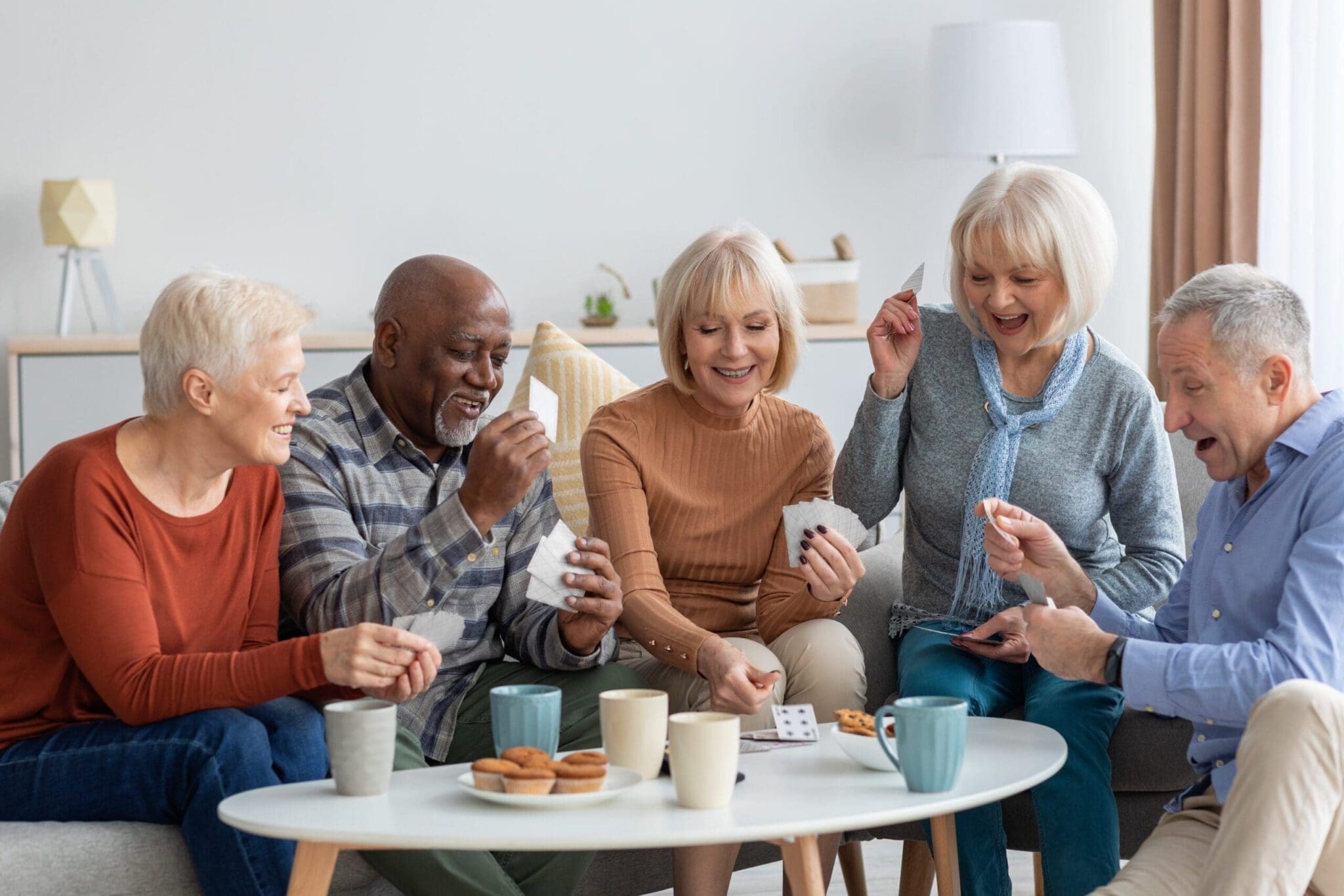 A group of senior friends sitting together on a cozy couch, playing cards and laughing. The setting includes warm lighting and coffee cups, highlighting the joy of social engagement in a retirement community.