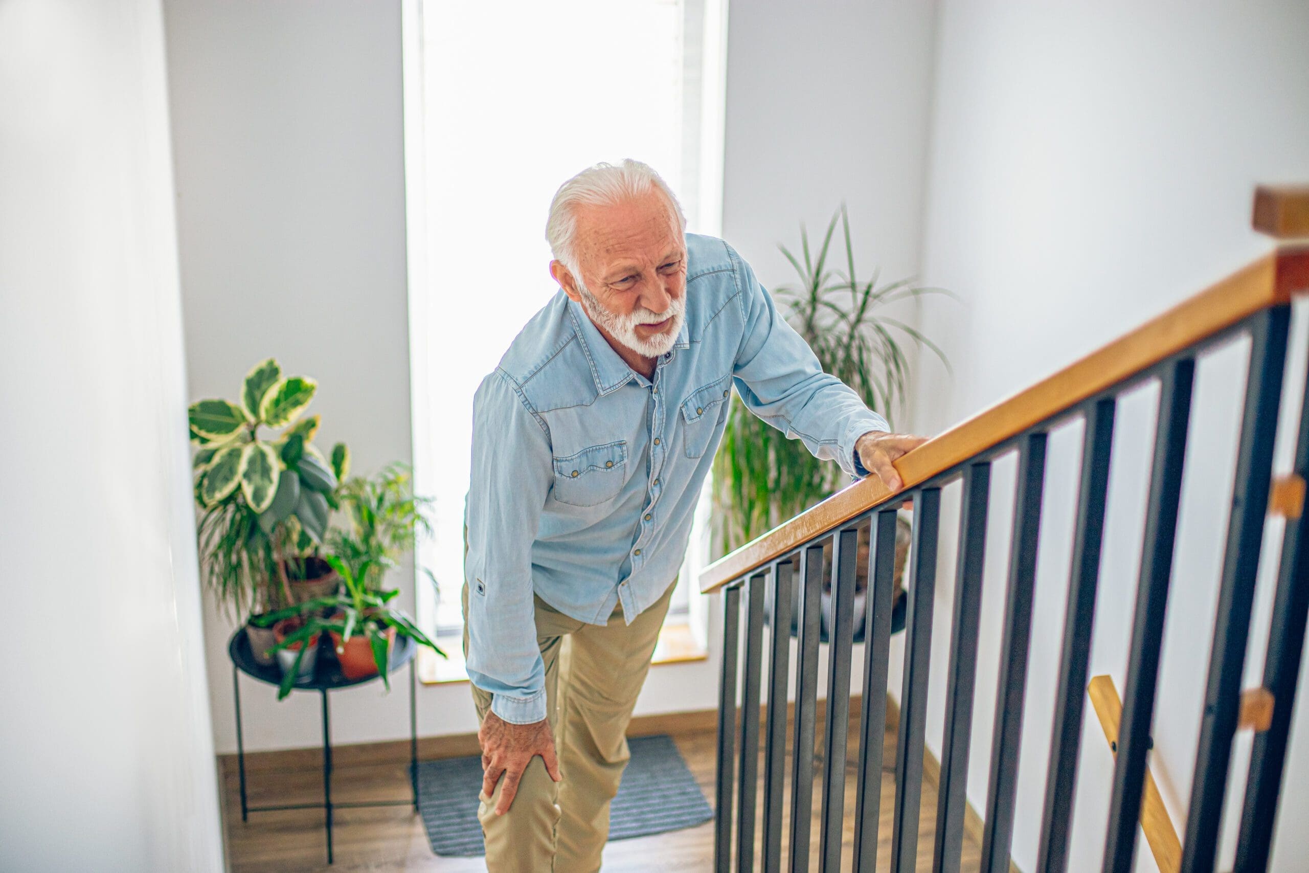 An elderly man with white hair and a beard grips a staircase railing while holding his knee, showing difficulty navigating stairs—a common safety concern for seniors aging at home.