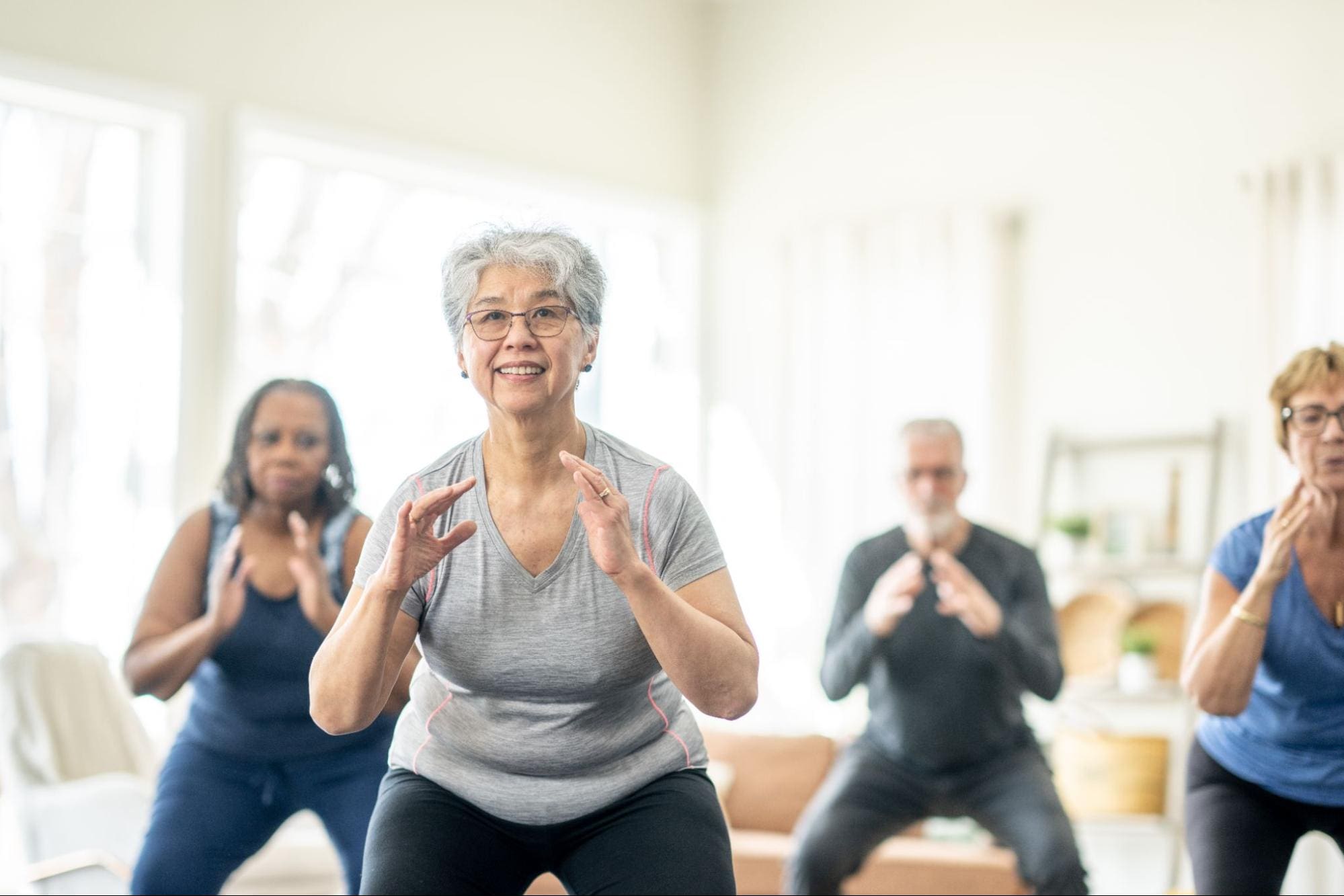 A group of seniors participating in a light fitness class in a bright, modern room. A smiling elderly woman in the foreground is engaged in a squat exercise, with other seniors following along behind her.