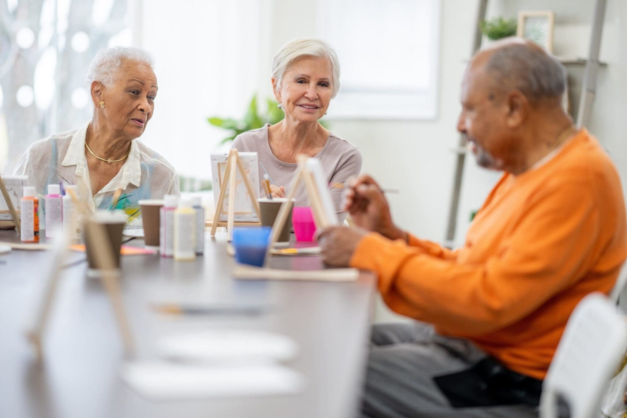 A group of seniors participating in an art class, painting on small canvases in a well-lit, creative space. They are smiling and interacting, highlighting the engaging activities available in a retirement community.