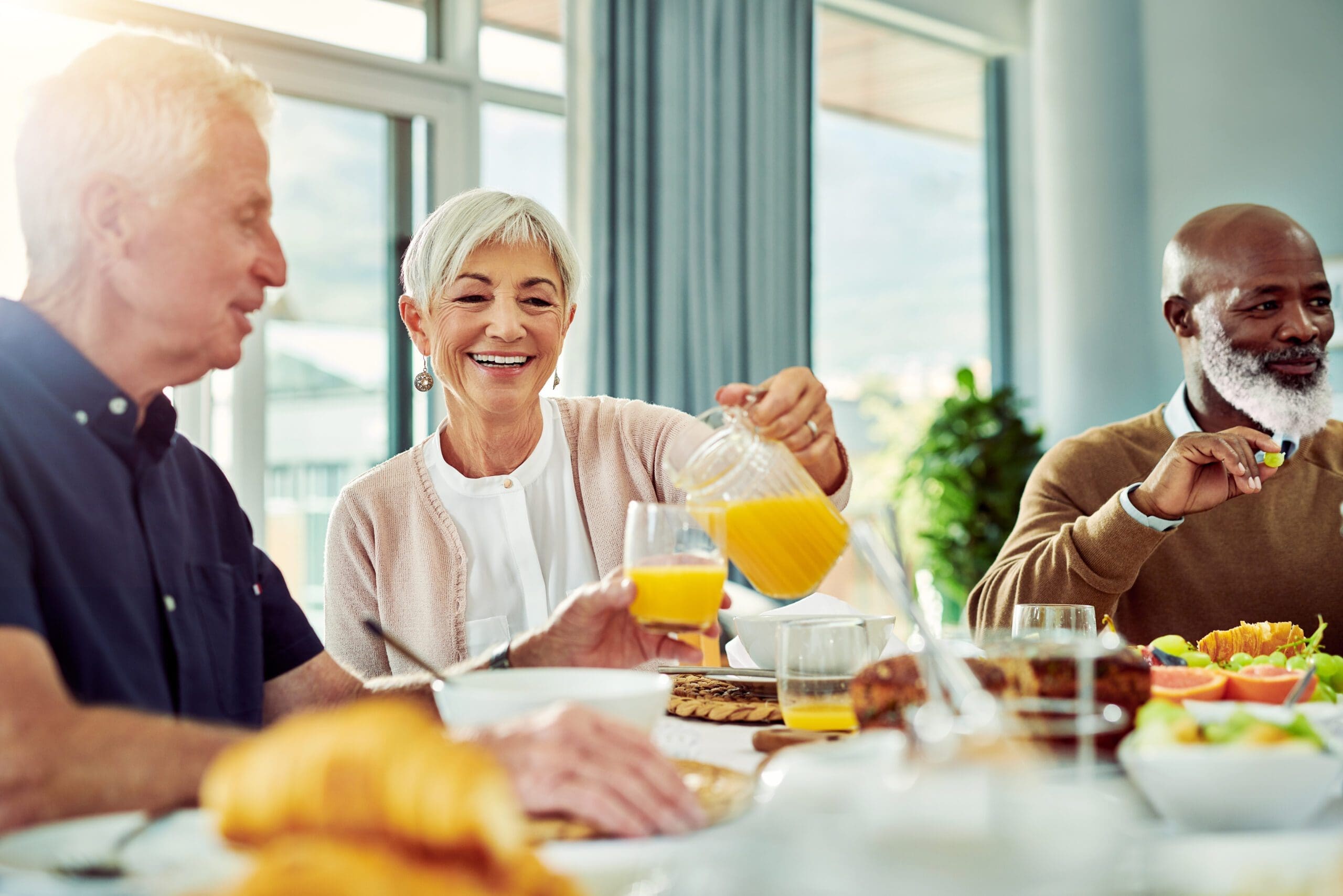 Smiling seniors sharing a meal at a retirement community, with one woman pouring orange juice and others enjoying food and conversation in a bright and cheerful dining space.