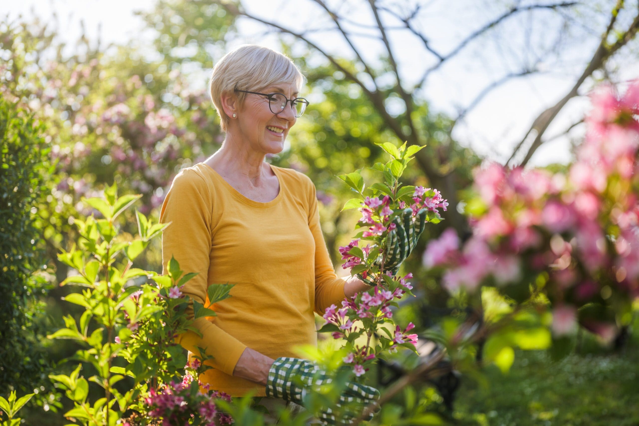 A smiling senior woman wearing a yellow shirt and gardening gloves, tending to blooming flowers in a vibrant and sunny outdoor garden at a retirement community.