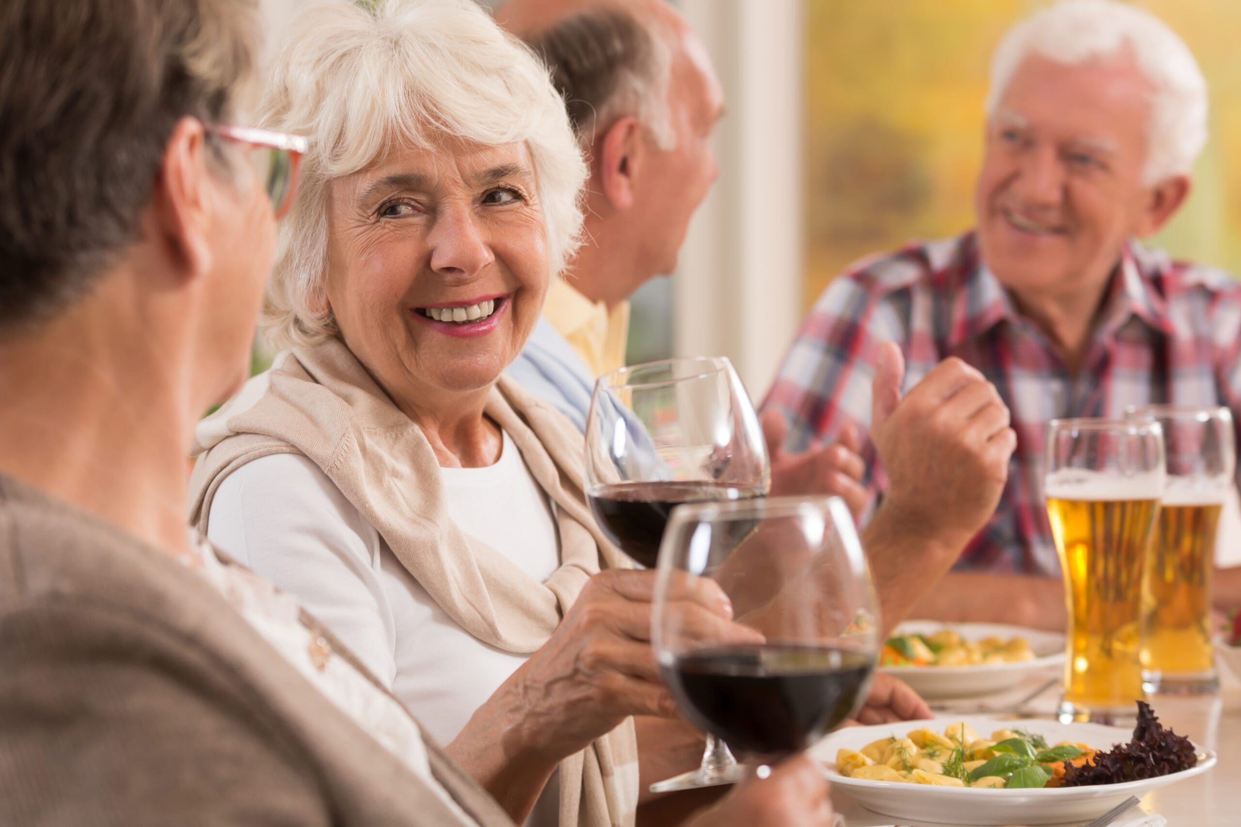 A group of seniors enjoying a social gathering over wine and a delicious meal at a well-decorated dining table, smiling and chatting warmly.