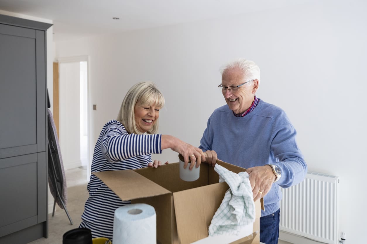 Senior couple smiling as they unpack household items from a cardboard box in their new retirement community.