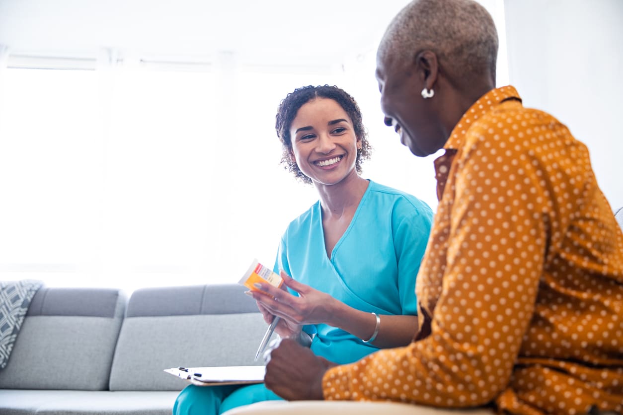 Alt Text: A friendly healthcare worker in scrubs speaks with a senior woman in her home about medication, both smiling during a consultation, symbolizing some of the services offered in many retirement residences.