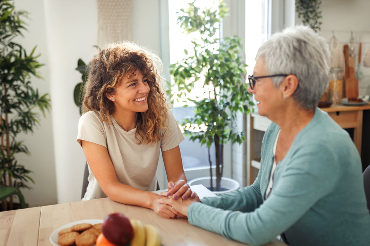 A senior woman smiles and holds hands with her adult daughter at a kitchen table, sharing a supportive conversation about moving into a retirement home.