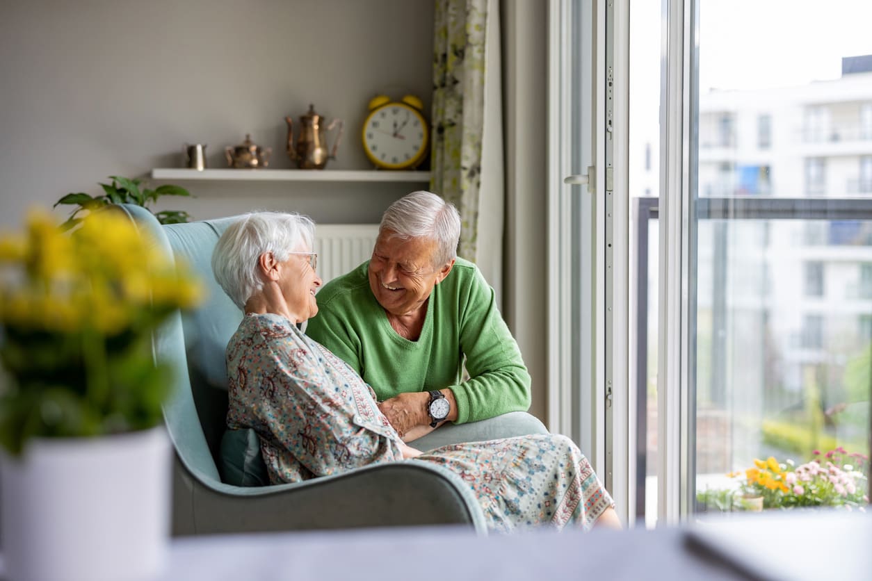 Senior couple sitting together and smiling in a comfortable retirement community suite.