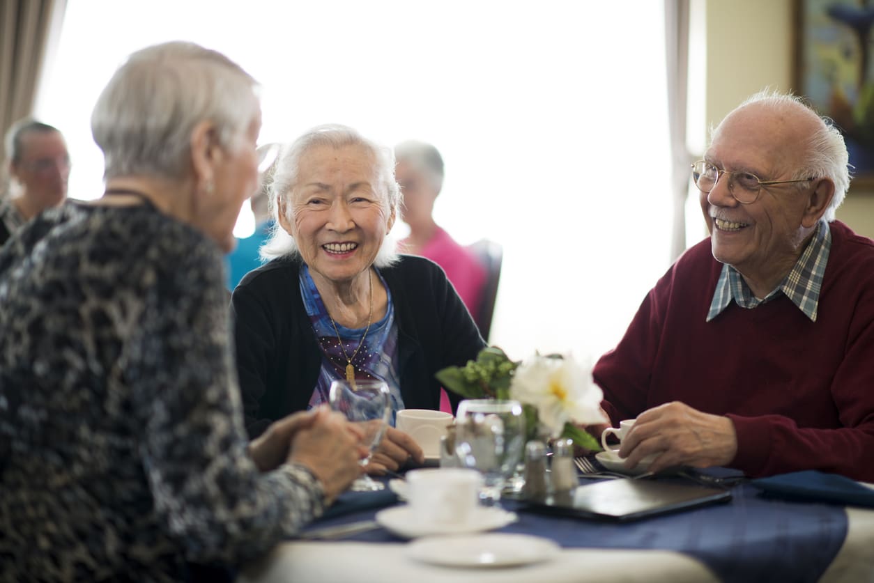 Group of older adults laughing and enjoying lunch together in a bright retirement community dining room.