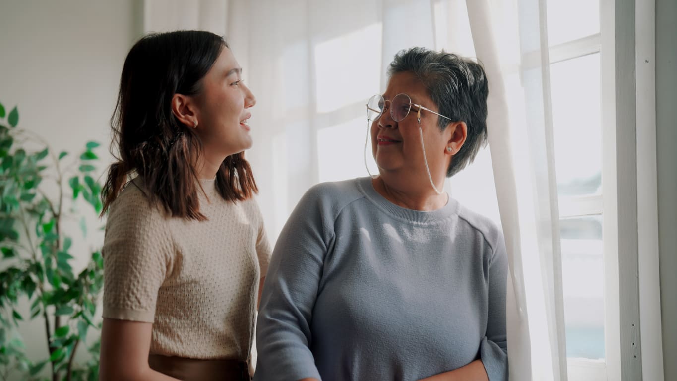 Daughter smiling at her mother inside a bright retirement community suite, sharing a warm, relaxed moment by the window.