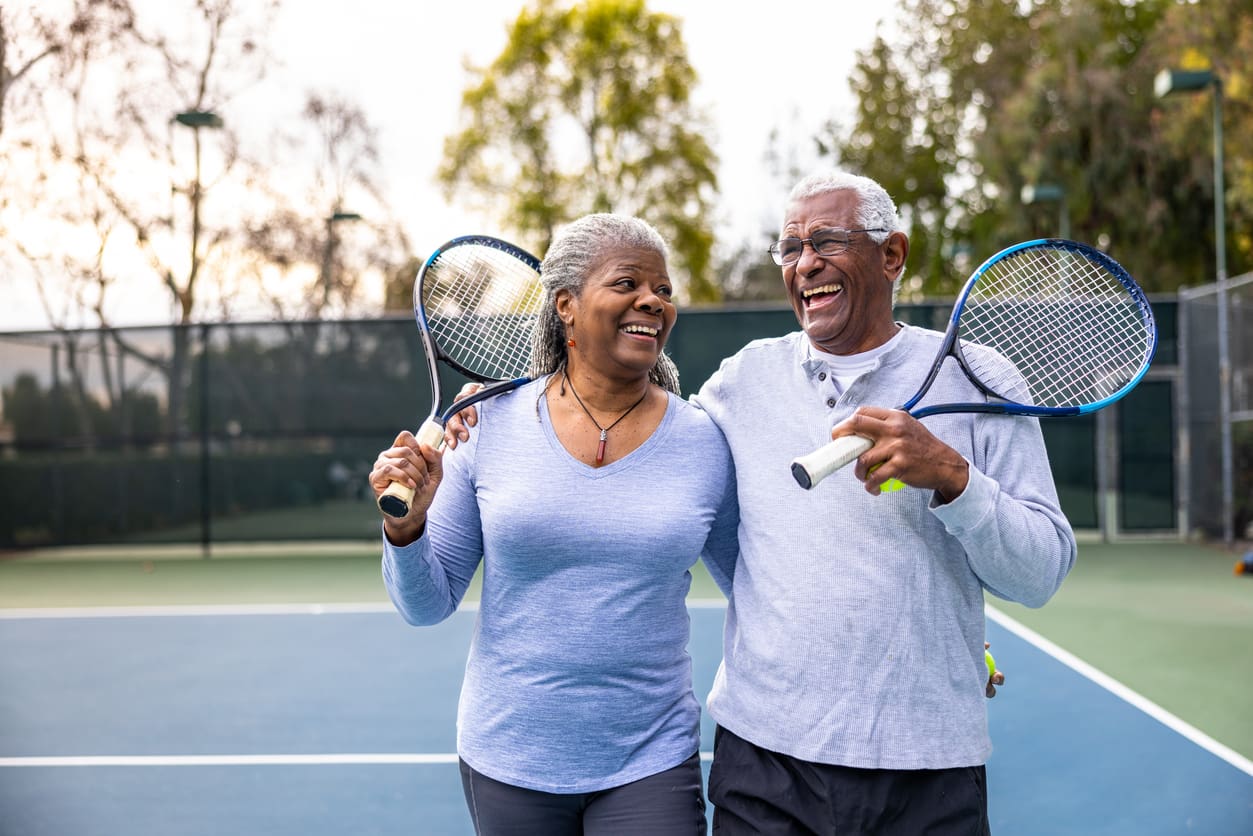 Senior couple laughing together after playing tennis outdoors, enjoying an active lifestyle as part of retirement living.